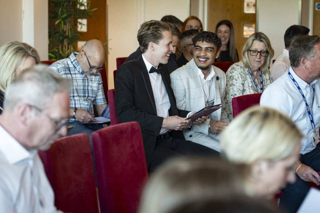 Two young men seated, one in a tuxedo smiling and looking at the other man who is also smiling, both holding papers. Other audience members are blurred in the background.