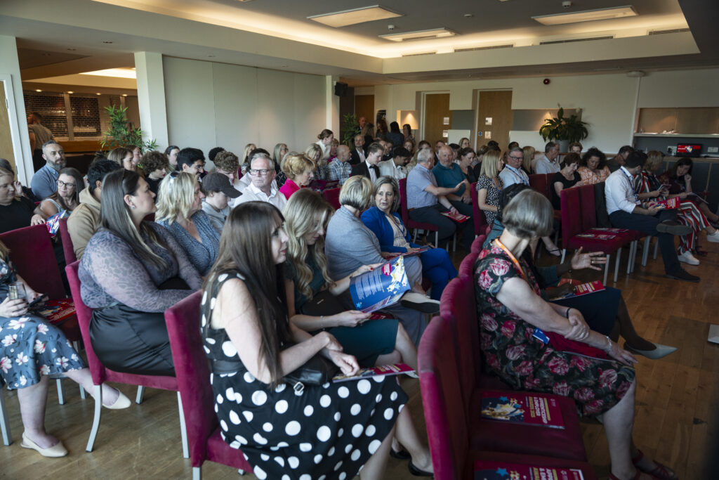 A wide shot of an audience seated on red chairs in a conference room, facing a stage or presentation area not visible in the frame.