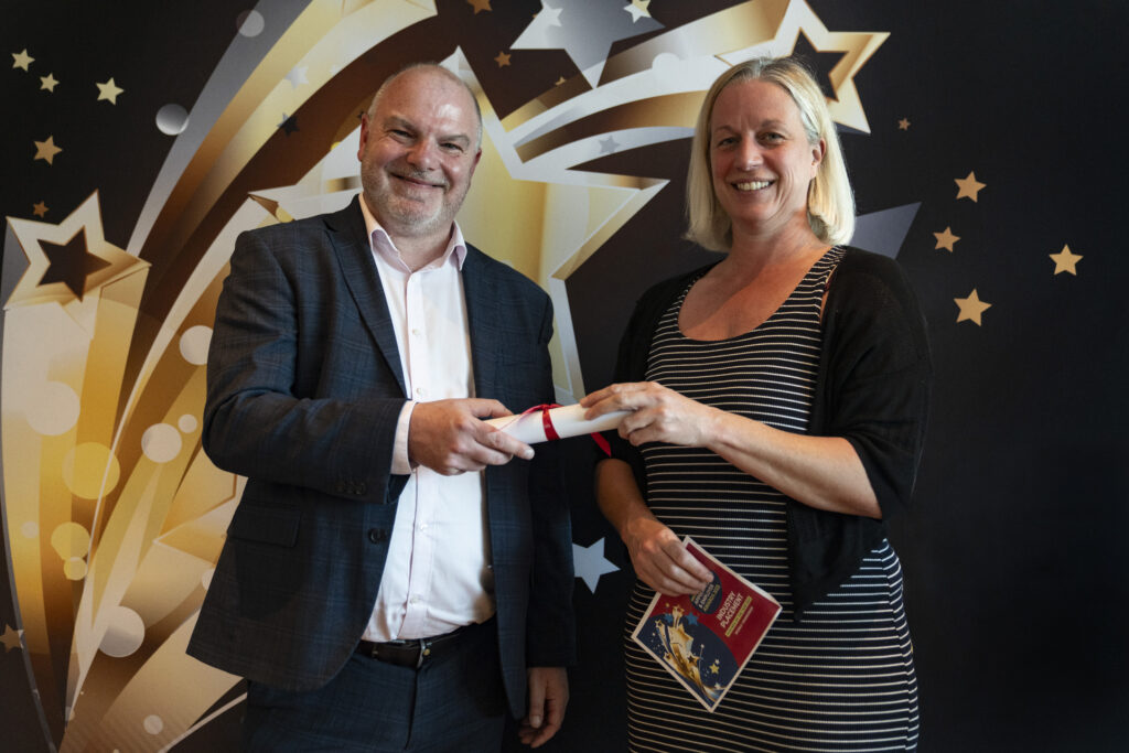 A smiling man in a suit and a smiling woman with blonde hair in a striped dress jointly hold a rolled-up certificate. The woman also holds an award booklet. They stand against a star-patterned background.