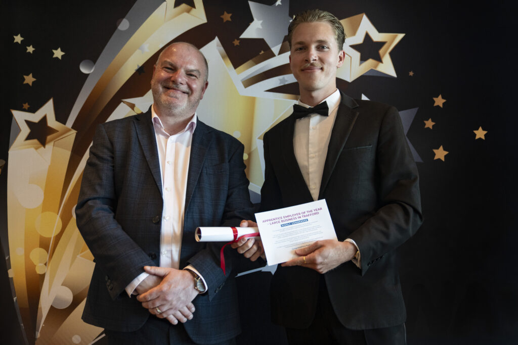 A smiling man in a suit and a smiling man in a tuxedo holding a certificate and paper, against a star-patterned background.