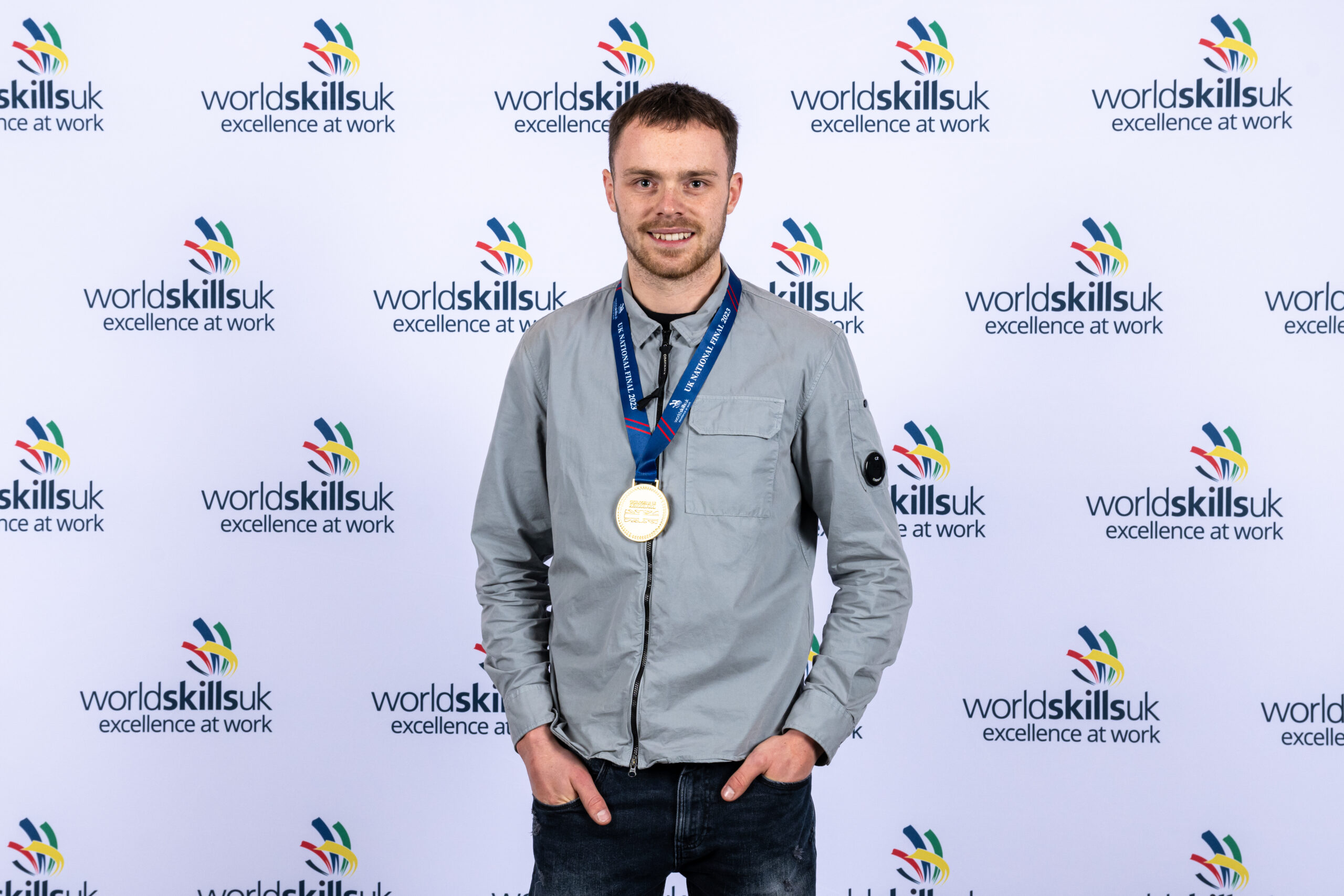 Adam Biddulph standing against a world skills background with a gold medal