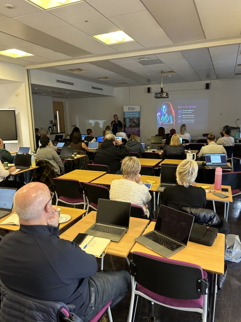 A classroom or conference room with attendees seated at tables using laptops, facing a projector screen displaying a colourful presentation.