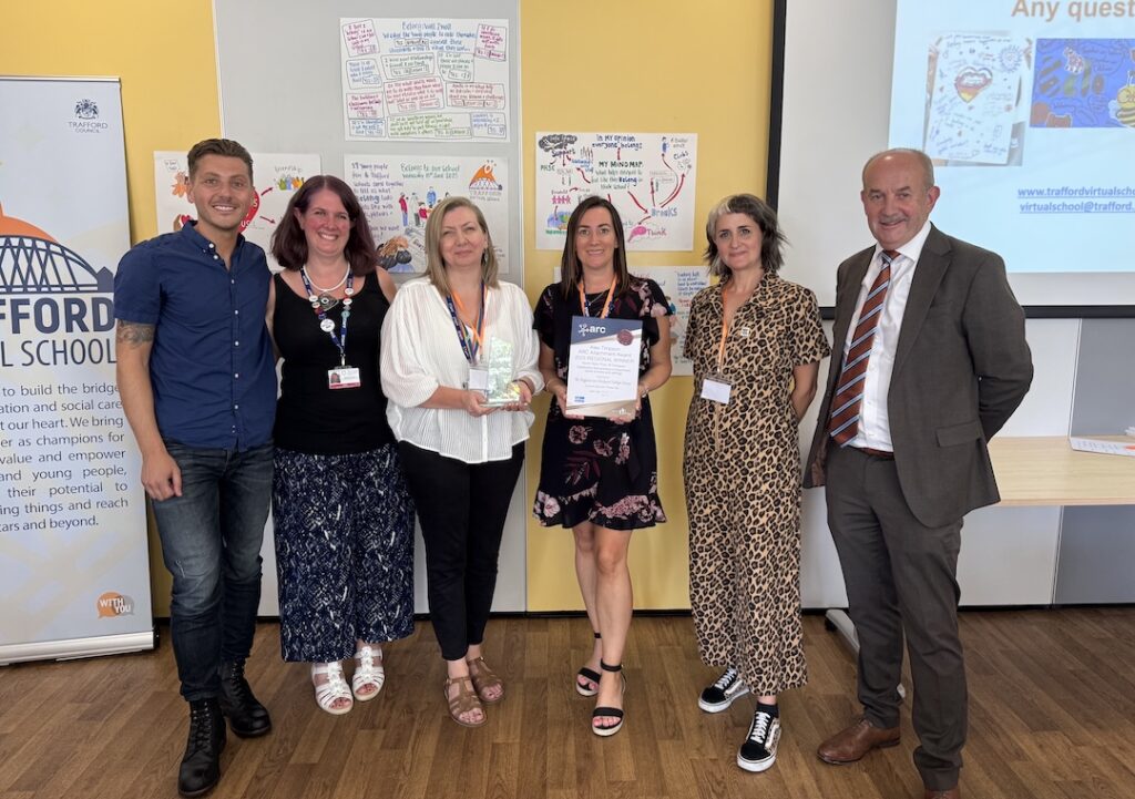 Team from Trafford posing with an award and certificate in front of a yellow and gray wall, with educational posters and a presentation screen in the background