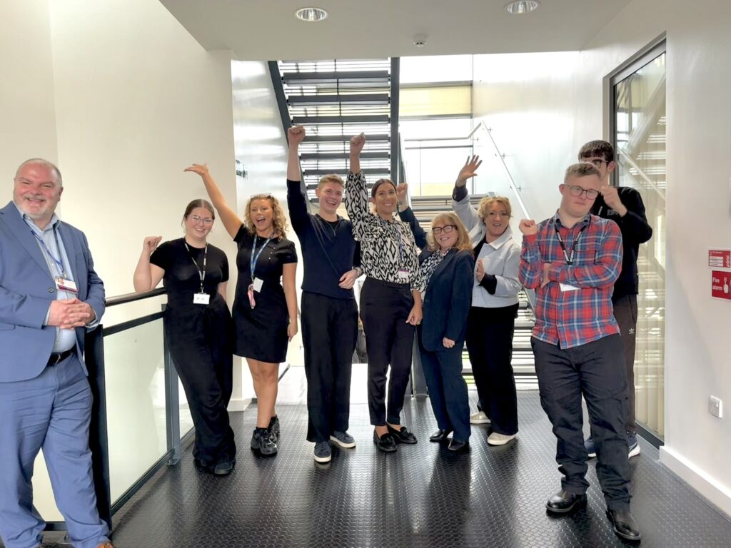Nine individuals posing energetically with raised arms and expressive gestures in a hallway setting