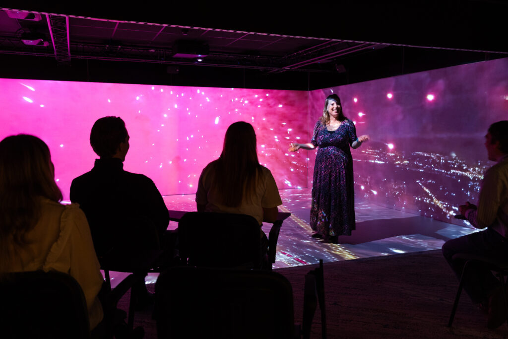 A woman stands and speaks in front of an immersive pink digital backdrop with bright lights and abstract visuals, addressing an audience of four seated people. The setting appears to be a modern presentation or workshop using projection or virtual reality technology to create an engaging visual environment.