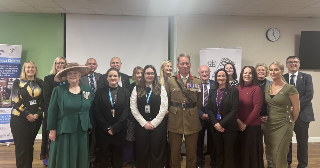 A group of people standing together indoors in front of two banners. The group includes individuals wearing formal attire, including one person in a military uniform with medals and another in a green outfit with a hat.