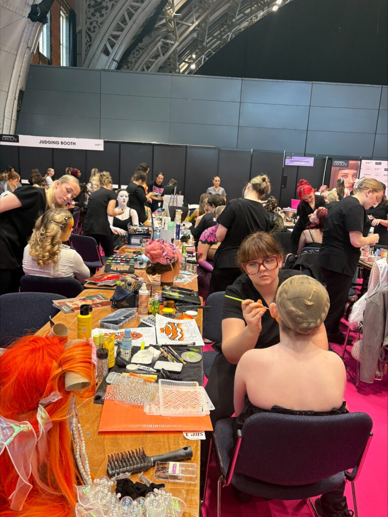 Busy makeup room with artists applying body paint and preparing wigs and tools at workstations.