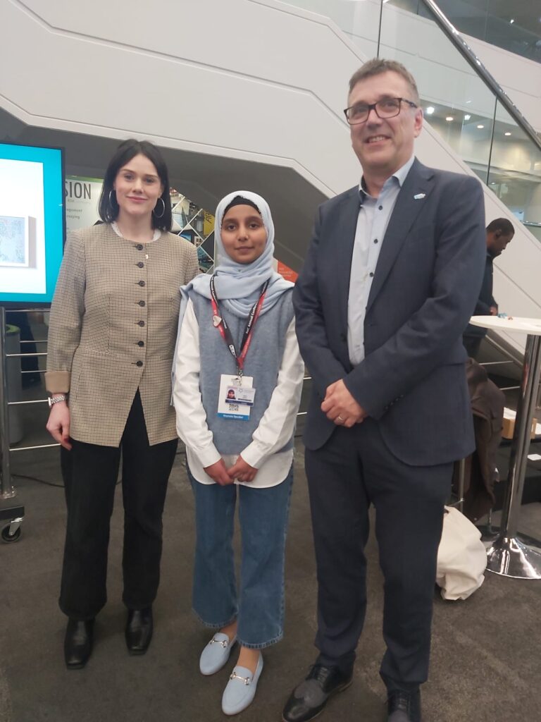 Hajira Qaiser at a college conference, standing with two professionals in a modern venue with presentation screens and networking space.