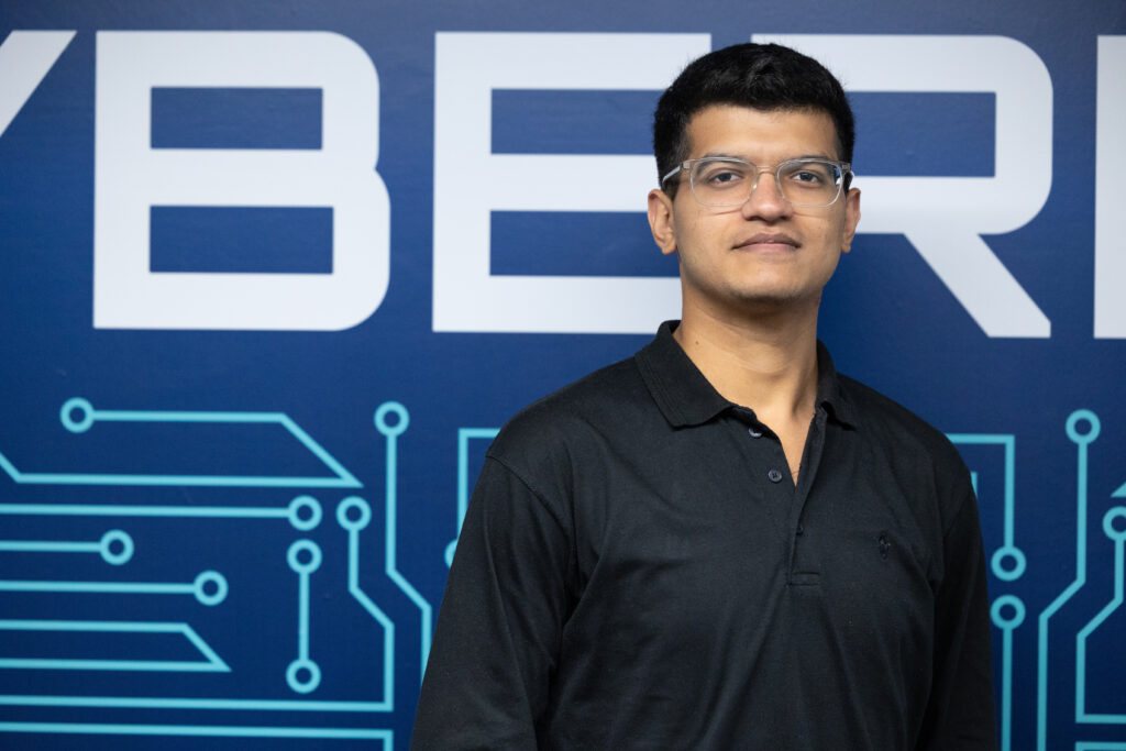 Hammad Jabeen standing in front of a blue wall with circuit board graphics and the word ‘CYBER’ visible, representing a cybersecurity learning environment