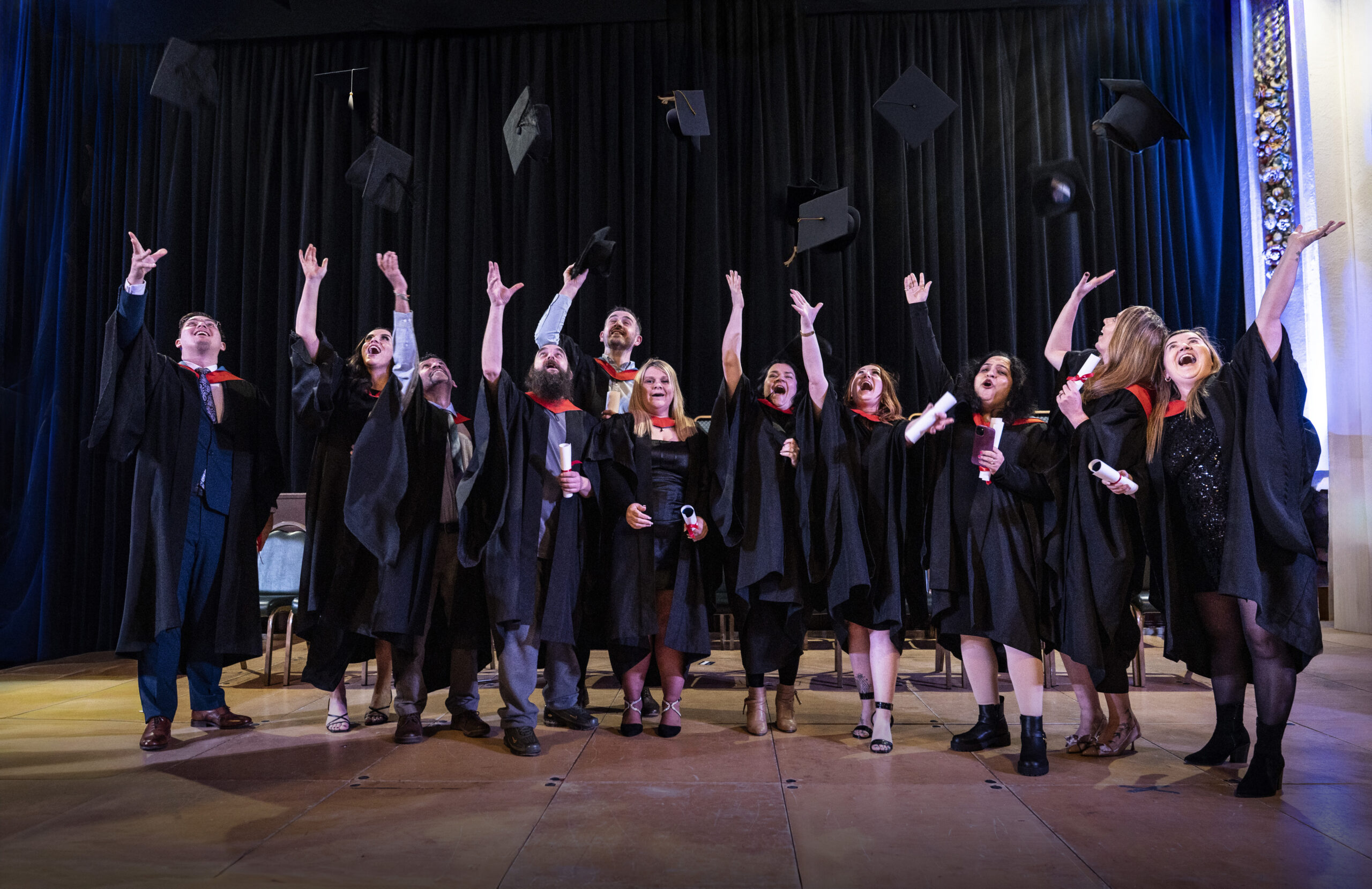 Graduates in black gowns and red stoles celebrate by throwing mortarboards into the air on stage during the university graduation ceremony at University Centre Stockport College.
