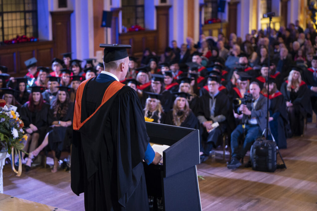 Academic speaker in graduation gown addresses graduates and guests from a podium in a large hall during the university graduation ceremony at University Centre Stockport College.