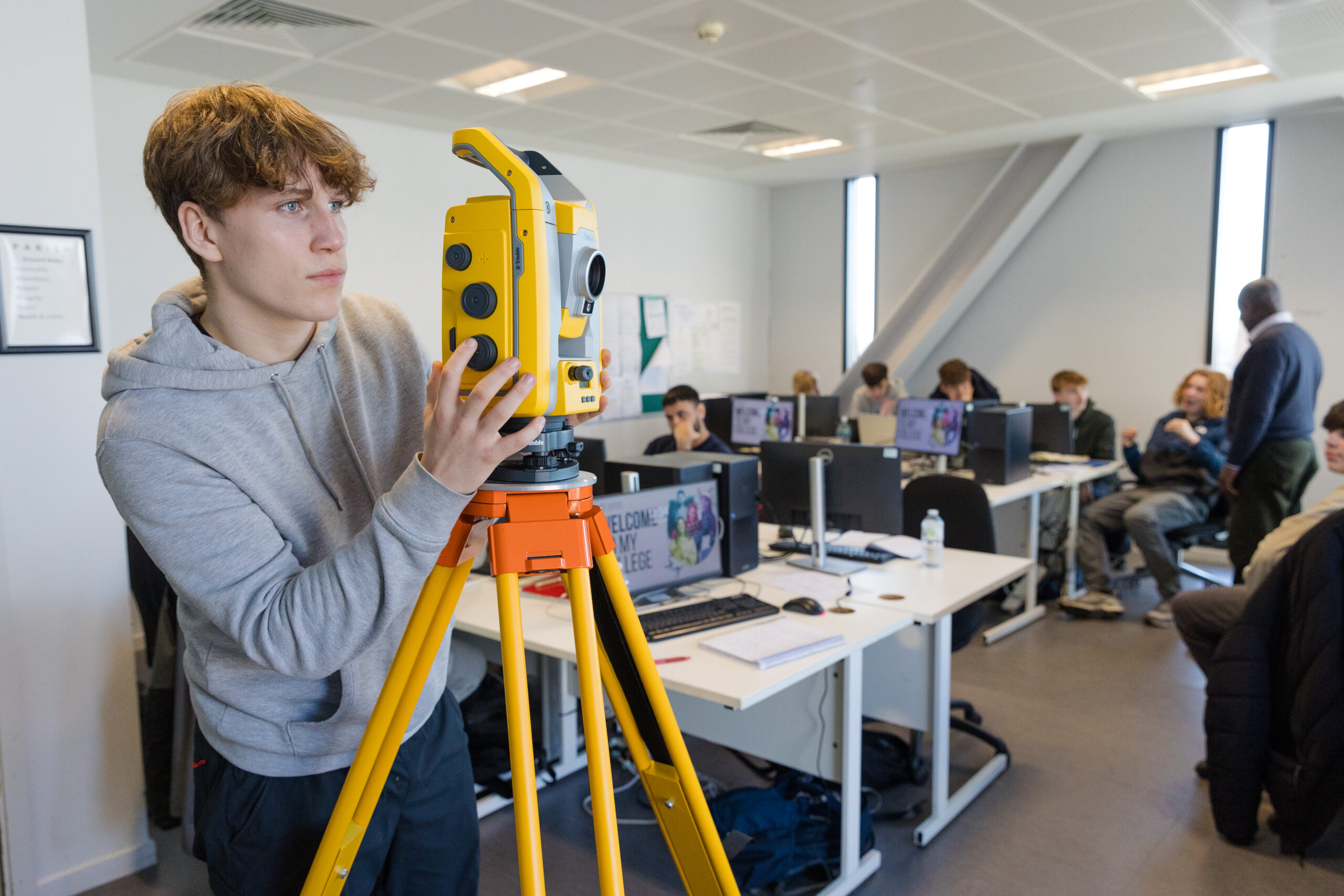 A young man stands at the front of a classroom where others sit behind computer monitors. The standing man is looking through a brigh yellow site surveying camera on a tripod.