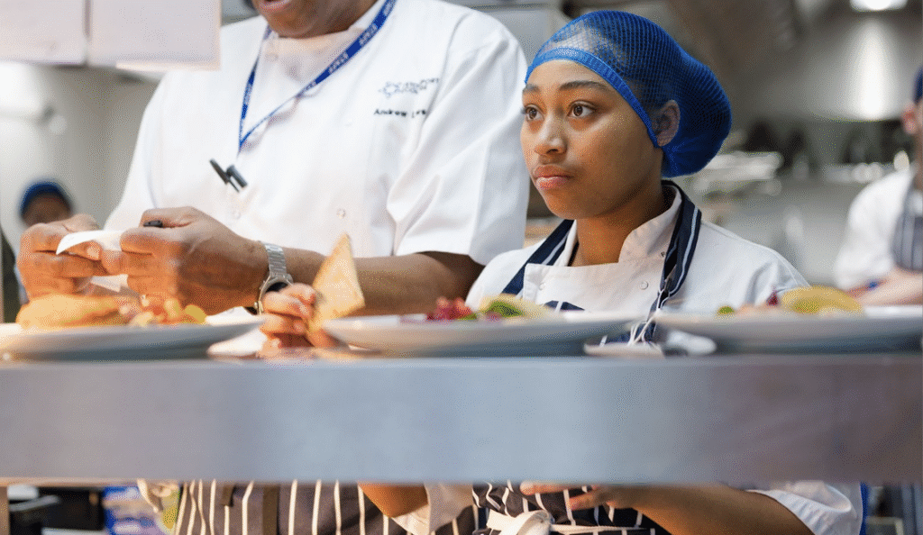 A young woman is stood in a professional kitchen wearing chef's clothing and a blue hairnet. She is in front of a service area, which is full of plated up food.